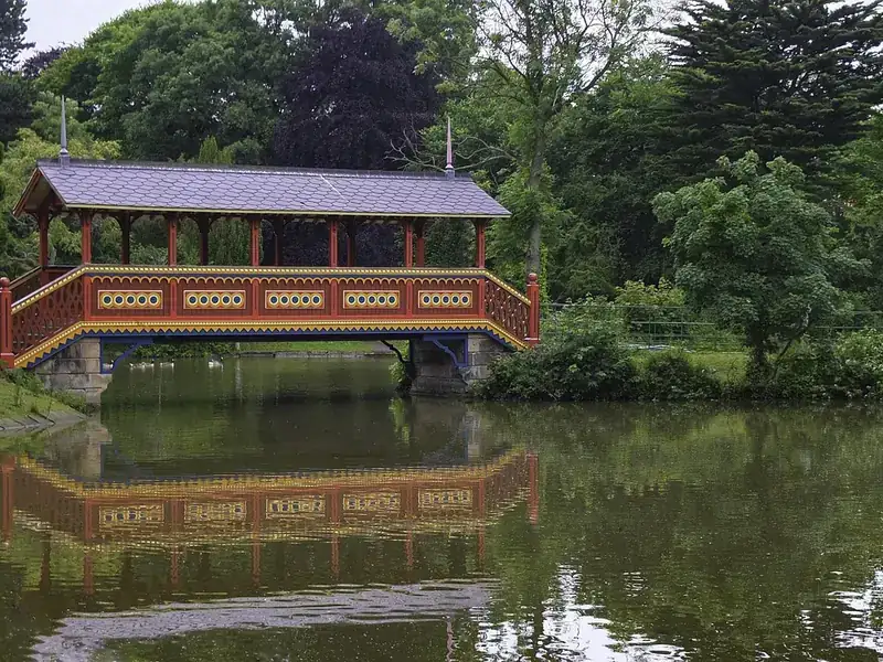 The ornate covered Swiss Bridge reflected in the lake in Birkenhead Park, surrounded by trees