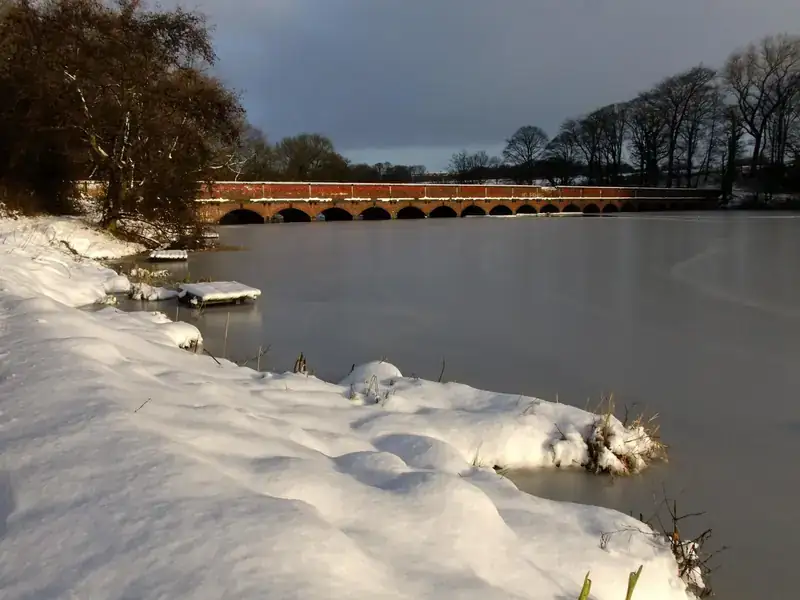 Carr Mill Dam in St Helens on a winter day, with snow on the bank and a long brick viaduct across the water