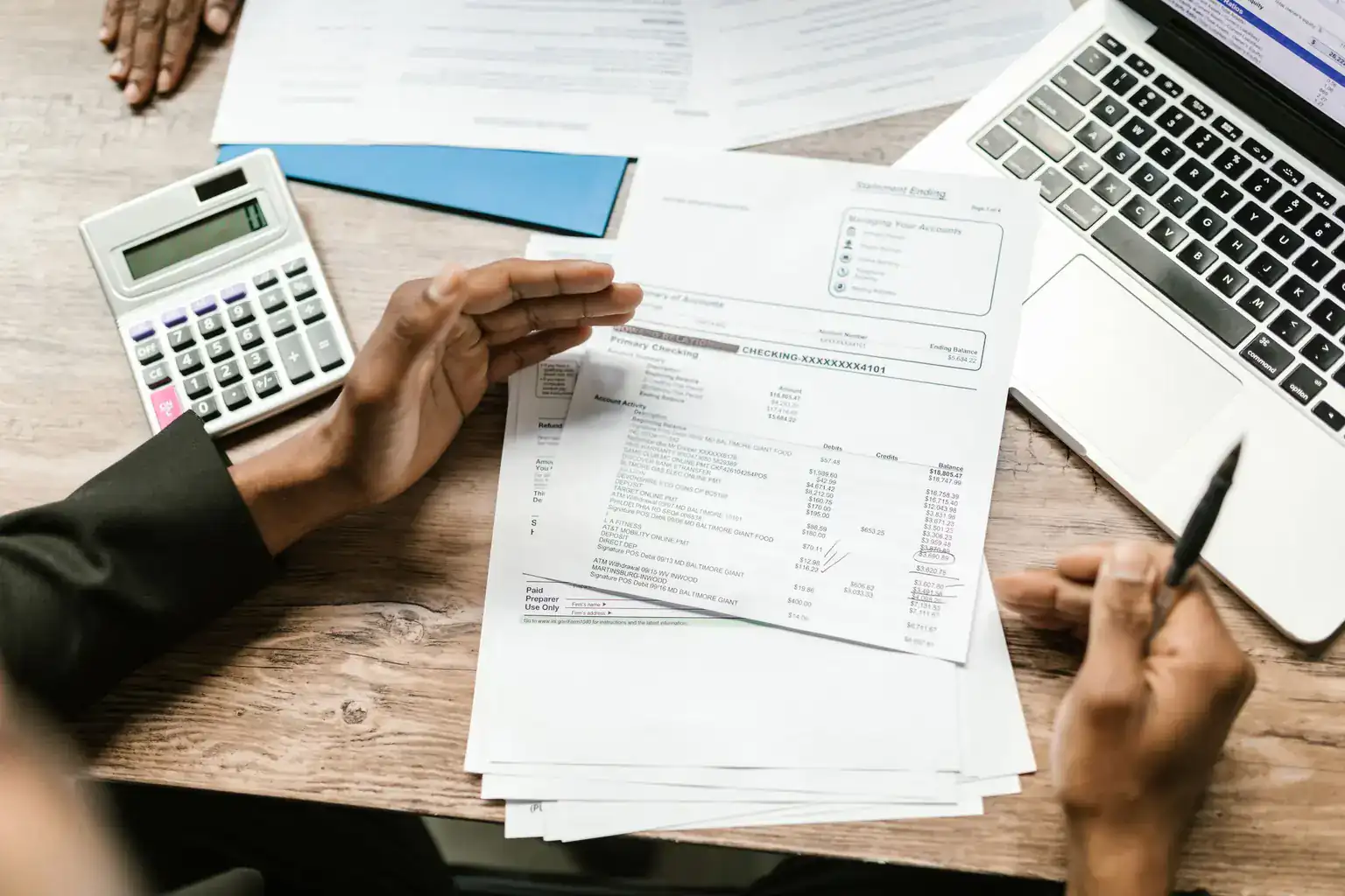 Hands spread out printed statements beside a calculator and laptop on a wooden desk