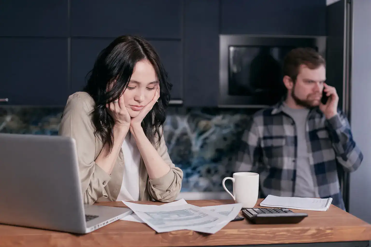 A worried woman looks over bills at the kitchen table as her partner makes a call