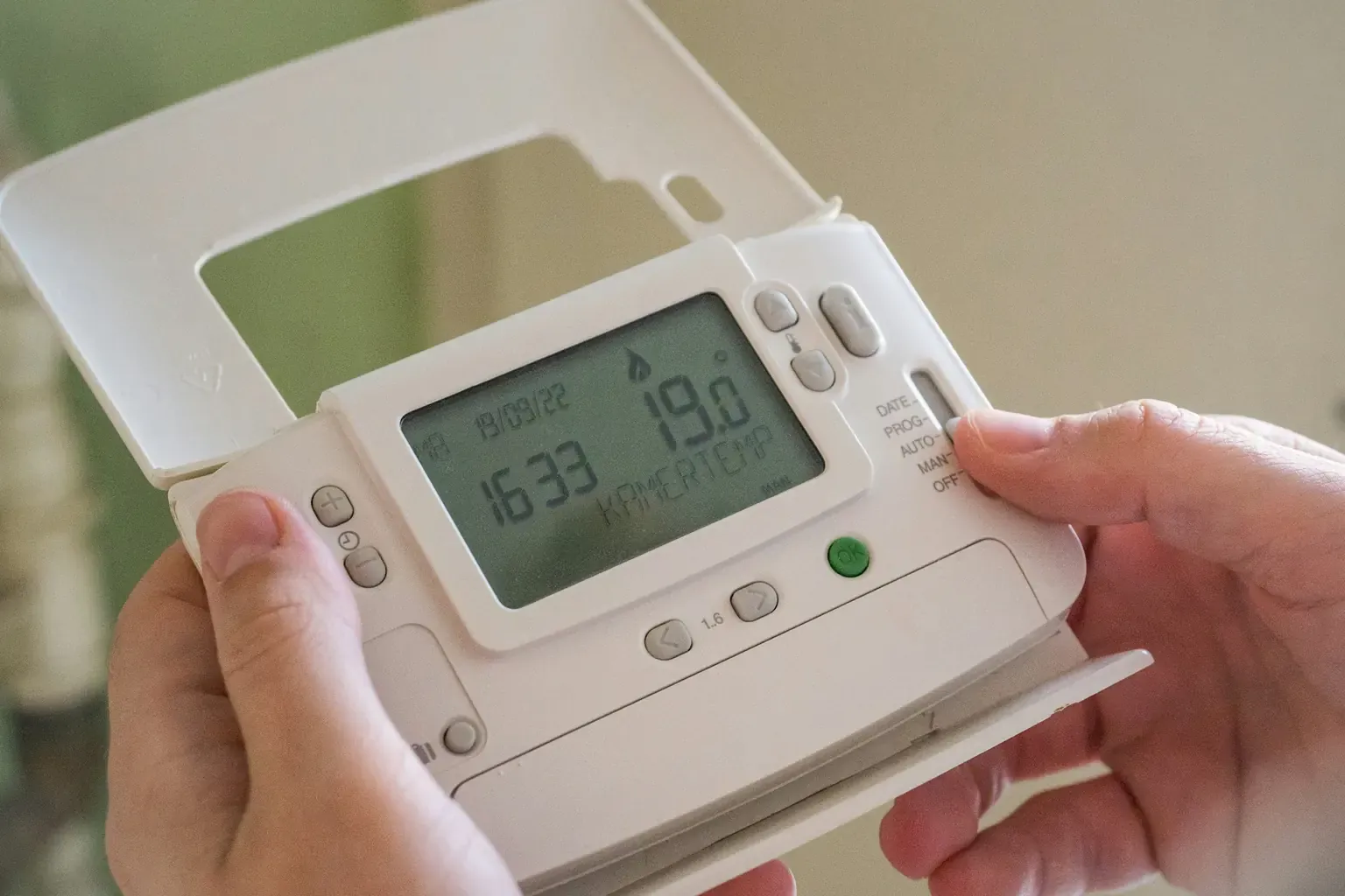 Hands holding a wall-mounted programmable heating thermostat showing the time and 19 degrees