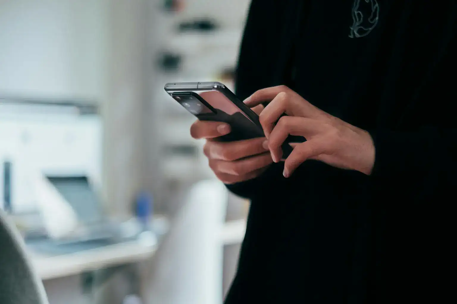 A man in a dark jumper scrolls on his smartphone in a bright, blurred room