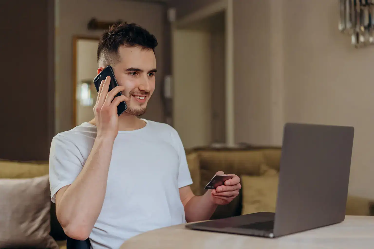 A young man at home talks on the phone while holding a bank card in front of his laptop