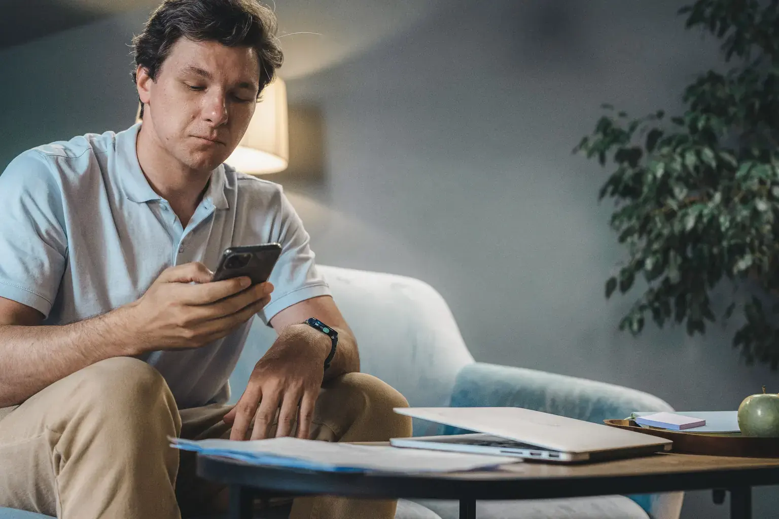 A man on a sofa checks his phone with paperwork and a laptop on the coffee table