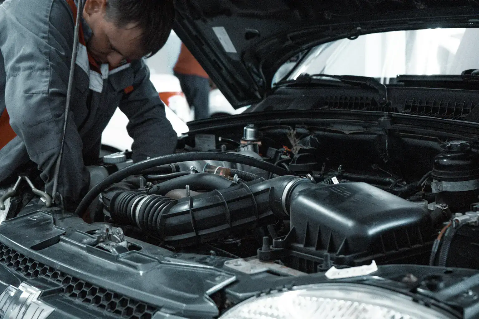 A mechanic in overalls leans over an open bonnet to inspect a car engine