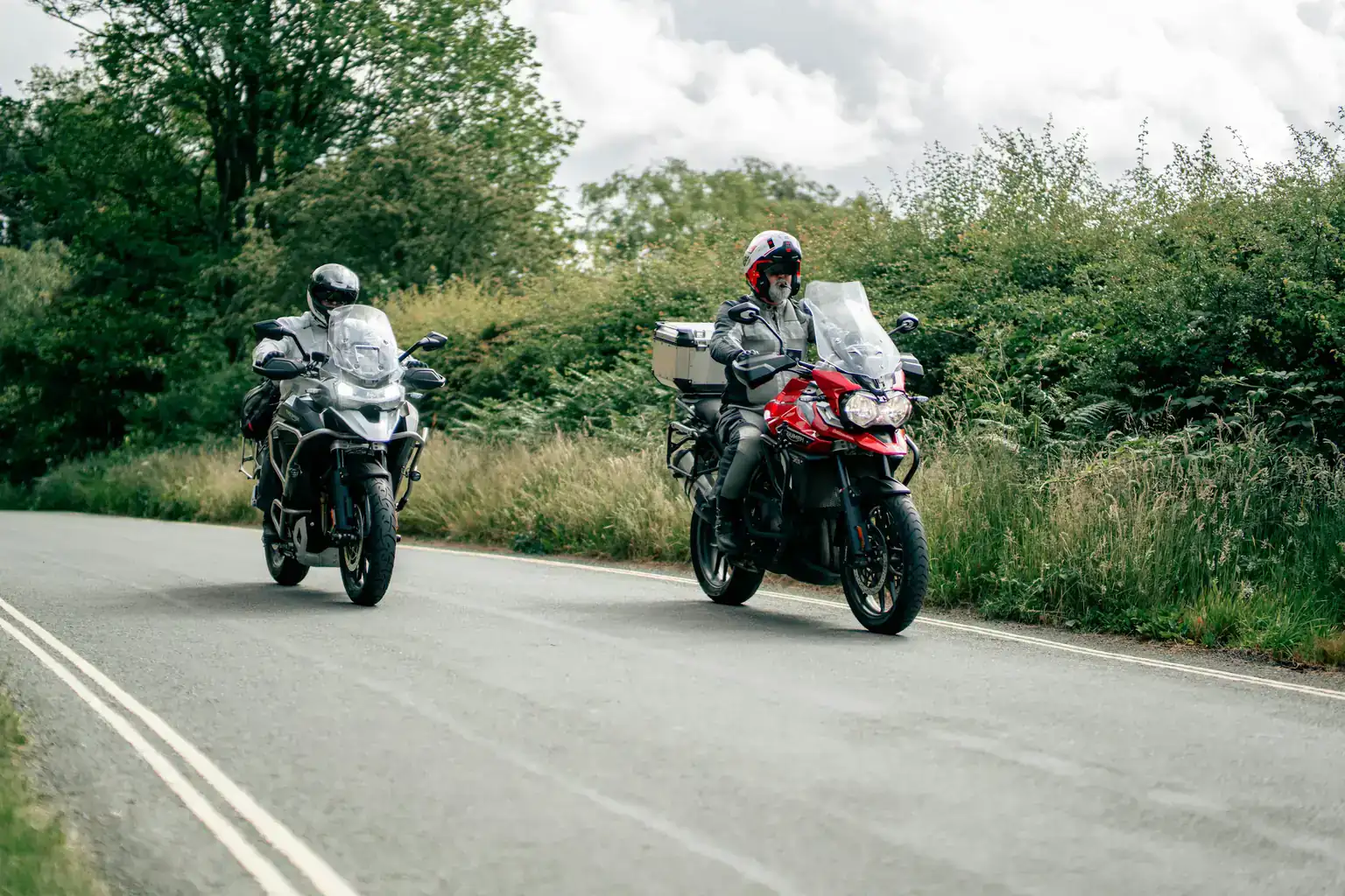 Two motorcyclists on touring bikes riding along a winding country road lined with hedges