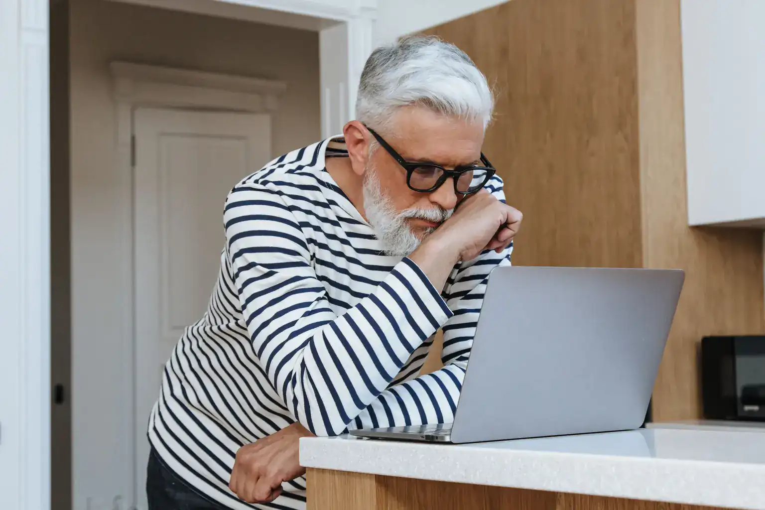 An older man in glasses leans on the worktop, studying his laptop screen closely