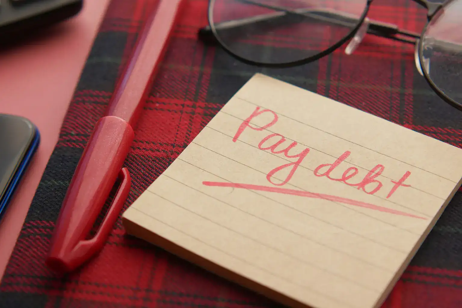 A sticky note reading pay debt beside a red pen and reading glasses