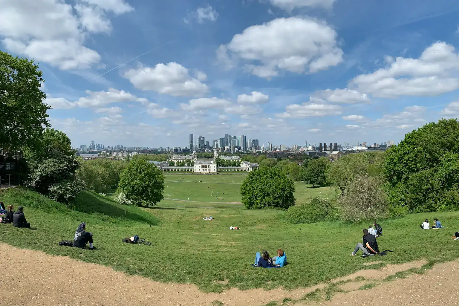 People sitting on the grass in Greenwich Park on a sunny day, with the London skyline beyond