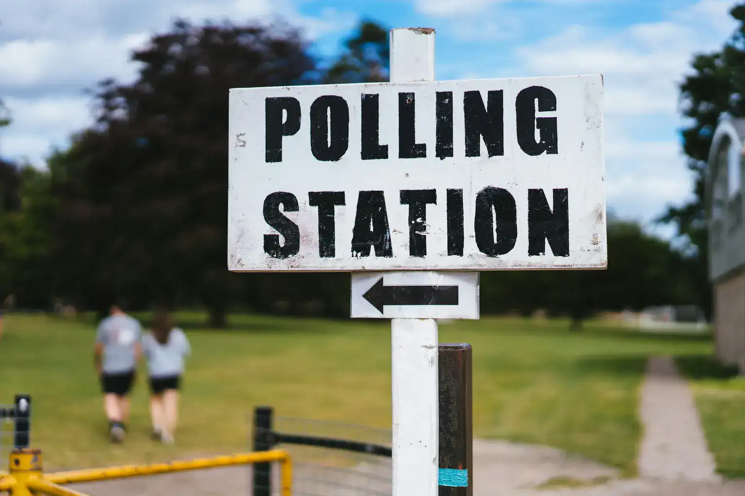 A white Polling Station sign with an arrow beside a park path on a sunny day
