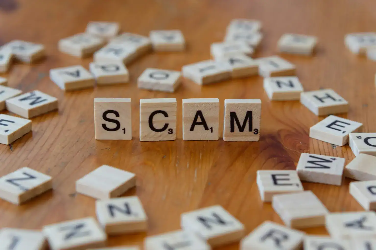 Wooden letter tiles spelling the word scam on a table among scattered tiles