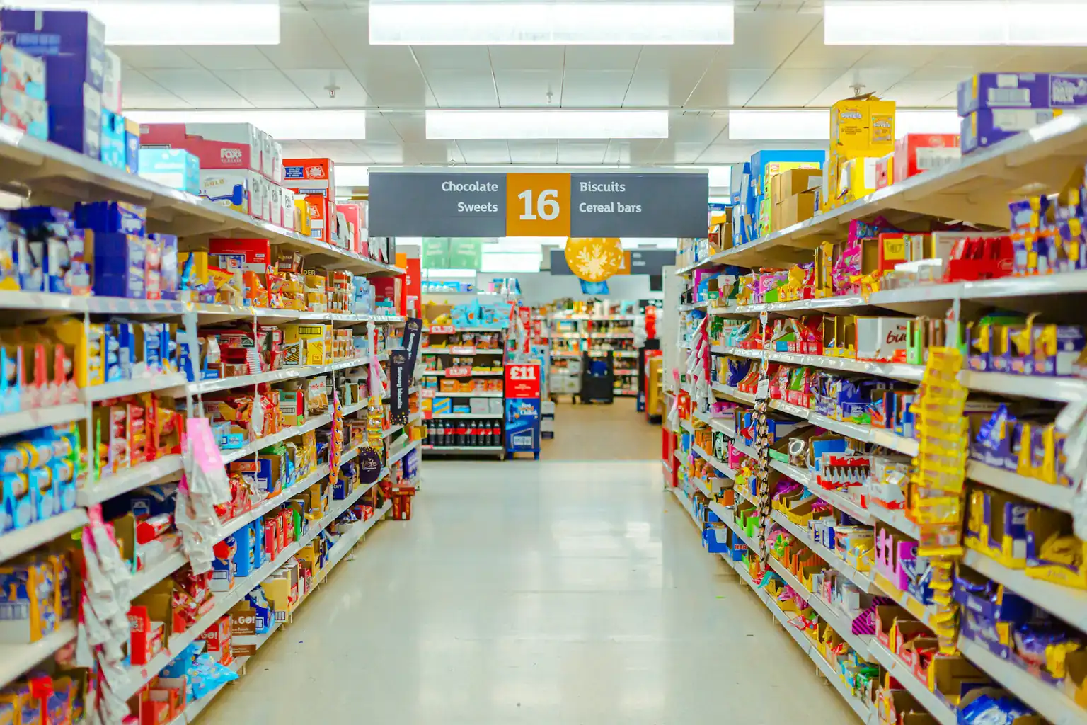 A long supermarket aisle with shelves of biscuits, sweets and cereal bars under bright lights