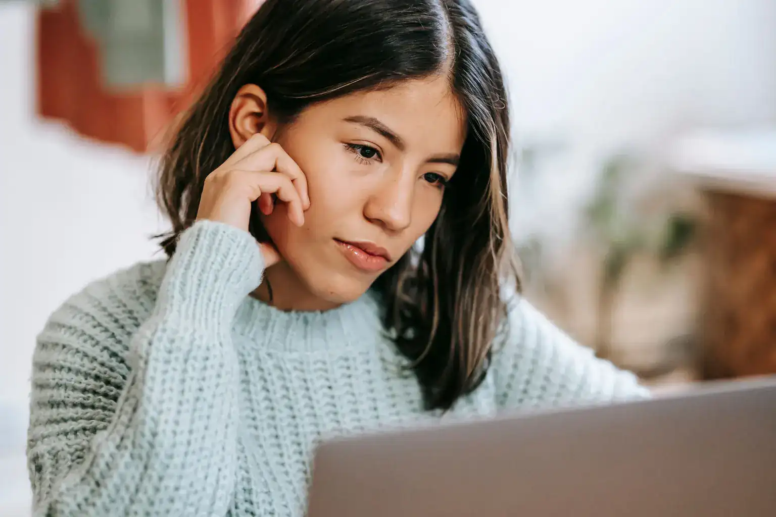 A young woman in a knitted jumper looks thoughtfully at her laptop at home