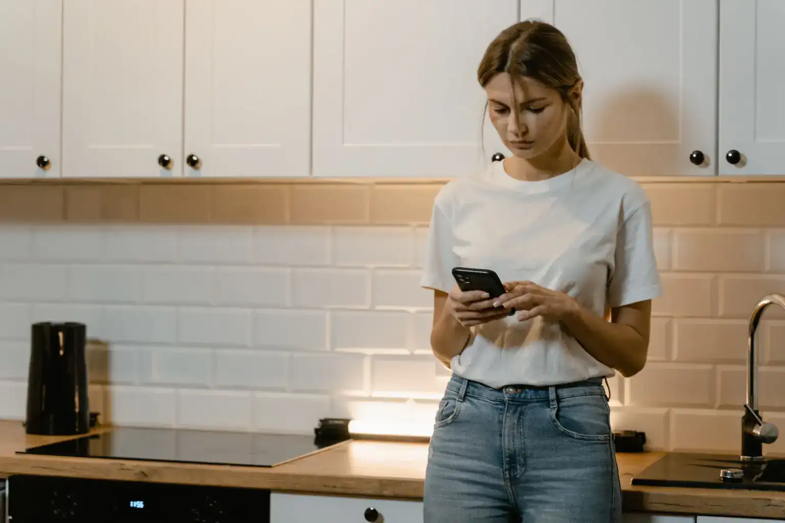 A young woman stands in her kitchen looking closely at her phone