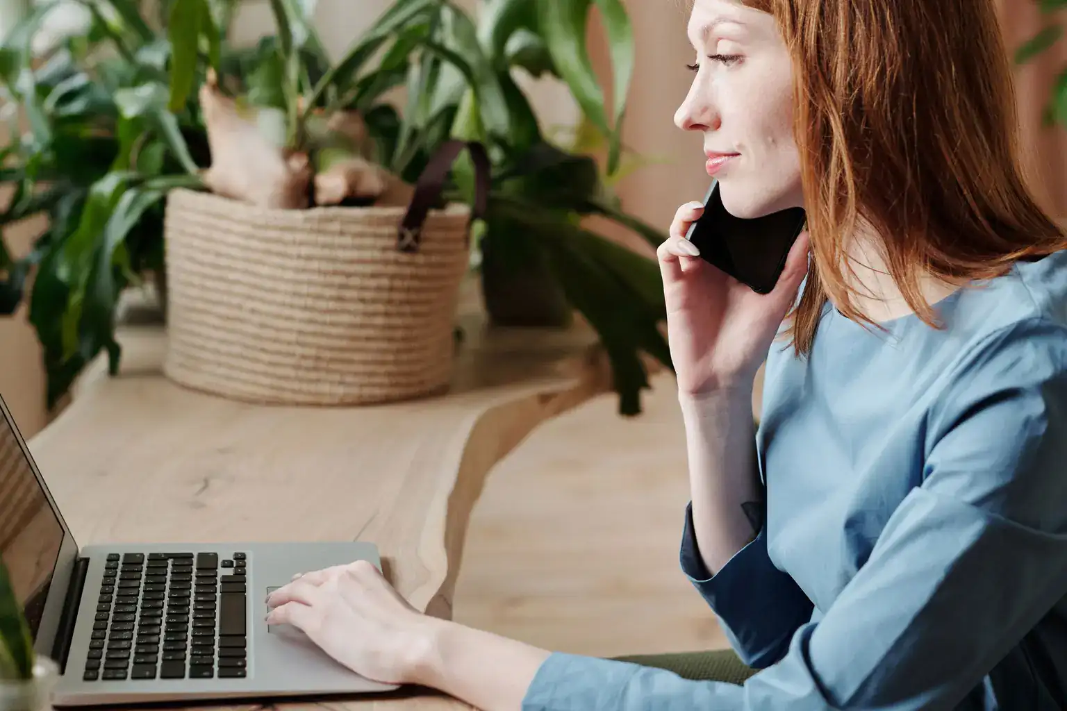 A woman with red hair makes a phone call while typing on a laptop by houseplants