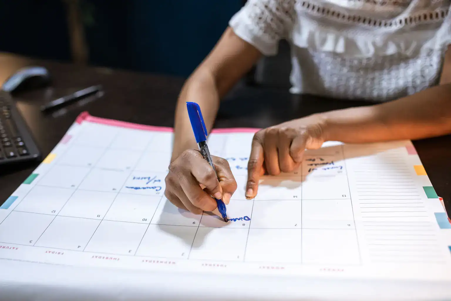 A woman writes notes on a large desk planner calendar with a blue pen