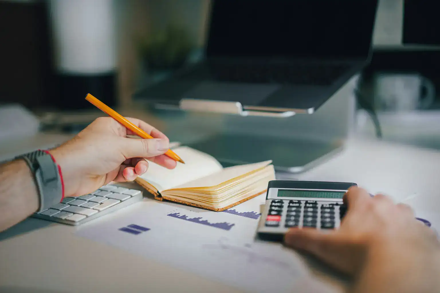 Hands using a calculator and pencil next to a notebook and printed charts on a desk