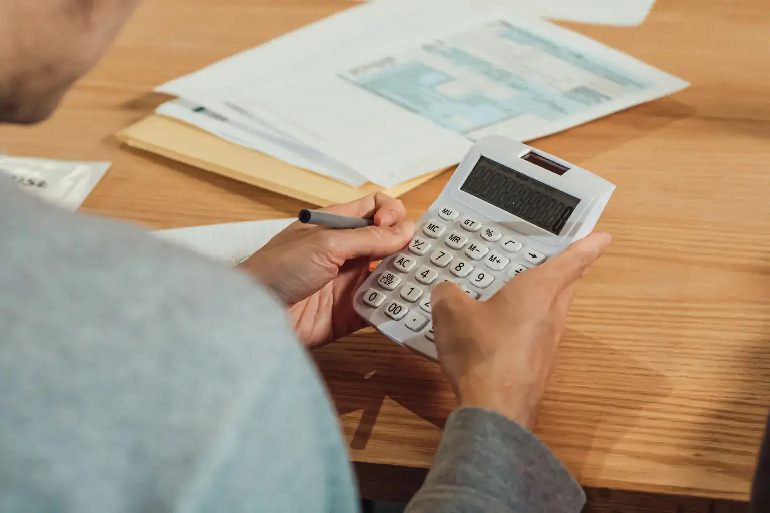 A man works out figures on a calculator with a pen beside a pile of paperwork