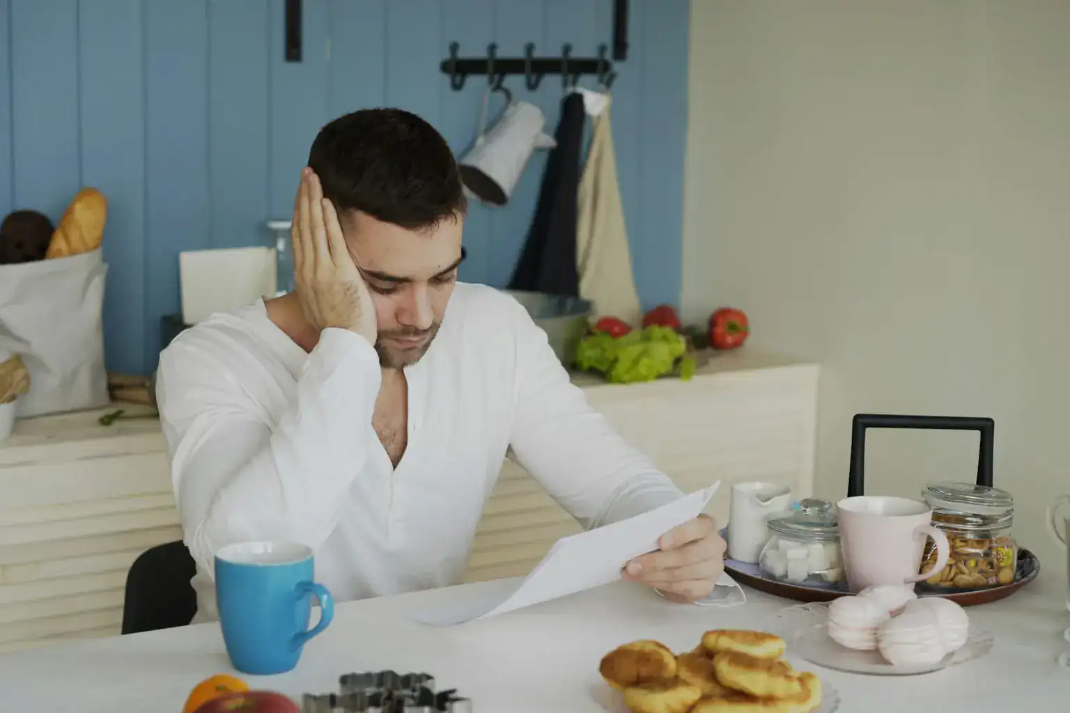 A worried young man holds his head while reading a letter at the kitchen table